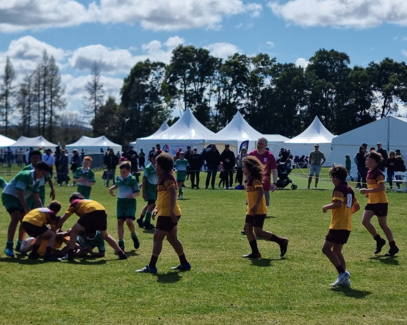 Children in yellow or green uniforms play rugby on a grassy field. The sun is shining and in the background are marquees. 